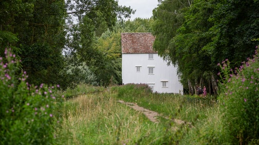 A view along a river towards a white clapboard mill building. It is summer and the foliage along the riverbank is lush and green